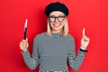 Beautiful blonde woman wearing french look with beret holding painter brushes smiling with an idea or question pointing finger Royalty Free Stock Photo