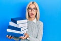 Beautiful blonde woman wearing glasses and holding pile of books afraid and shocked with surprise and amazed expression, fear and Royalty Free Stock Photo