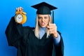 Beautiful blonde woman wearing graduation cap and ceremony robe holding alarm clock annoyed and frustrated shouting with anger,