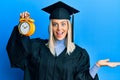 Beautiful blonde woman wearing graduation cap and ceremony robe holding alarm clock celebrating victory with happy smile and