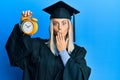 Beautiful blonde woman wearing graduation cap and ceremony robe holding alarm clock covering mouth with hand, shocked and afraid