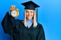 Beautiful blonde woman wearing graduation cap and ceremony robe holding alarm clock looking positive and happy standing and