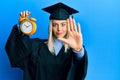 Beautiful blonde woman wearing graduation cap and ceremony robe holding alarm clock with open hand doing stop sign with serious