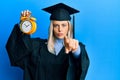 Beautiful blonde woman wearing graduation cap and ceremony robe holding alarm clock pointing with finger to the camera and to you,