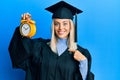 Beautiful blonde woman wearing graduation cap and ceremony robe holding alarm clock pointing finger to one self smiling happy and