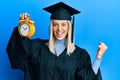 Beautiful blonde woman wearing graduation cap and ceremony robe holding alarm clock screaming proud, celebrating victory and