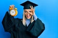 Beautiful blonde woman wearing graduation cap and ceremony robe holding alarm clock smiling happy doing ok sign with hand on eye