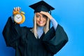 Beautiful blonde woman wearing graduation cap and ceremony robe holding alarm clock stressed and frustrated with hand on head,