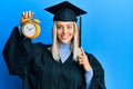 Beautiful blonde woman wearing graduation cap and ceremony robe holding alarm clock surprised with an idea or question pointing