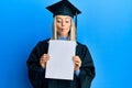 Beautiful blonde woman wearing graduation cap and ceremony robe holding blank banner making fish face with mouth and squinting