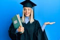 Beautiful blonde woman wearing graduation cap and ceremony robe holding books celebrating victory with happy smile and winner Royalty Free Stock Photo