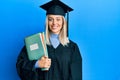 Beautiful blonde woman wearing graduation cap and ceremony robe holding books looking positive and happy standing and smiling with Royalty Free Stock Photo