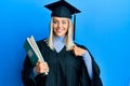 Beautiful blonde woman wearing graduation cap and ceremony robe holding books pointing finger to one self smiling happy and proud Royalty Free Stock Photo