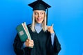 Beautiful blonde woman wearing graduation cap and ceremony robe holding books pointing thumb up to the side smiling happy with Royalty Free Stock Photo