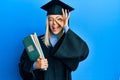 Beautiful blonde woman wearing graduation cap and ceremony robe holding books smiling happy doing ok sign with hand on eye looking Royalty Free Stock Photo