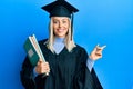 Beautiful blonde woman wearing graduation cap and ceremony robe holding books smiling happy pointing with hand and finger to the Royalty Free Stock Photo