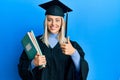 Beautiful blonde woman wearing graduation cap and ceremony robe holding books smiling happy and positive, thumb up doing excellent Royalty Free Stock Photo