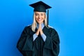 Beautiful blonde woman wearing graduation cap and ceremony robe praying with hands together asking for forgiveness smiling