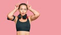 Beautiful blonde woman wearing gym clothes and using headphones doing funny gesture with finger over head as bull horns