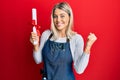 Beautiful blonde woman wearing hairdresser apron holding degree screaming proud, celebrating victory and success very excited with Royalty Free Stock Photo