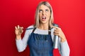 Beautiful blonde woman wearing hairdresser apron and holding scissors angry and mad screaming frustrated and furious, shouting Royalty Free Stock Photo