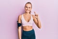 Beautiful blonde woman wearing sportswear and arm band smiling and confident gesturing with hand doing small size sign with Royalty Free Stock Photo