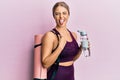 Beautiful blonde woman wearing sportswear holding water bottle and yoga mat sticking tongue out happy with funny expression Royalty Free Stock Photo