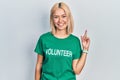 Beautiful blonde woman wearing volunteer t shirt cheerful with a smile on face pointing with hand and finger up to the side with
