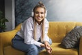 Beautiful blonde woman in white headphones sits on yellow sofa, enjoys listening to podcast. Relaxing girl in shirt