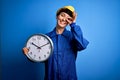 Beautiful blonde worker woman with blue eyes wearing security helmet holding big clock with happy face smiling doing ok sign with Royalty Free Stock Photo