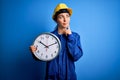 Beautiful blonde worker woman with blue eyes wearing security helmet holding big clock serious face thinking about question, very Royalty Free Stock Photo