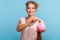 Beautiful blonde young adult woman in summer dress investing in bitcoins, putting golden crypto coin into piggy bank, looking at Royalty Free Stock Photo