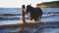 A beautiful blonde young woman standing next to a horse at a lake