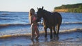 A beautiful blonde young woman standing next to a horse at a lake