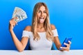 Beautiful blonde young woman holding dollars and using smarpthone sticking tongue out happy with funny expression Royalty Free Stock Photo