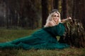 A beautiful blonde young woman in a long green dress and a diadem on her head in the forest. girl sitting near the old stump. Royalty Free Stock Photo