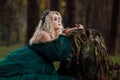 A beautiful blonde young woman in a long green dress and a diadem on her head in the forest. girl sitting near the old stump. Royalty Free Stock Photo