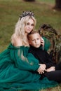 A beautiful blonde young woman in a long green dress and a diadem on her head with stylish young son in the forest. girl and boy Royalty Free Stock Photo