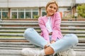 Beautiful blonde young woman smiling broadly, wearing blue jeans, white t-shirt and pink jacket, smiling and sitting on the bench Royalty Free Stock Photo