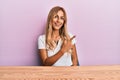 Beautiful blonde young woman wearing casual clothes sitting on the table cheerful with a smile of face pointing with hand and Royalty Free Stock Photo