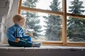 Beautiful blue-eyed baby sitting on the windowsill and looks out the window at the green trees Royalty Free Stock Photo