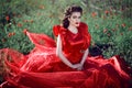 Beautiful blue-eyed young lady with perfect make up and hairstyle wearing luxurious silk red ball gown sitting in the poppy field