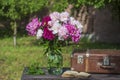 Beautiful bouquet of flowers with red, pink and white peonies in a glass jar with water and old suitcase, book on a wooden table Royalty Free Stock Photo