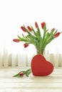 Beautiful bouquet of red tulips in a vase and red heart gift box on the white wooden table by the window with white curtains, Royalty Free Stock Photo