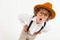 Beautiful boy with curly hair in white shirt, brown hat, glasses with black suspenders stands isolated on white