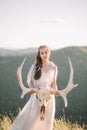 Beautiful bride brunette girl in white long dress is made of tulle looks down, keeps on hand a deer skull with horns decoration on Royalty Free Stock Photo