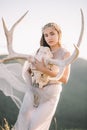 Beautiful bride brunette girl in white long dress is made of tulle looks down, keeps on hand a deer skull with horns decoration on Royalty Free Stock Photo