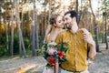 Beautiful bride embraces her groom by the shoulder. Wedding walk in a forest. Girl in white dress and man in an olive