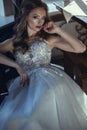 Beautiful bride in luxurious A-line wedding dress sitting in the vintage arm chair in relaxed pose