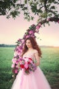 Beautiful bride in pink dress standing with her bouquet at a round flower arch under a large single tree in a field at sunset. Royalty Free Stock Photo